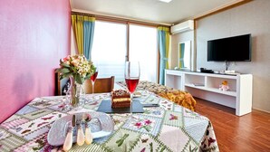 Habitación de hotel con una mesa de comedor preparada para dos, un jarrón de flores, una copa de vino tinto y una porción de pastel. Se observa un televisor en la pared y una estantería blanca con un oso de peluche.