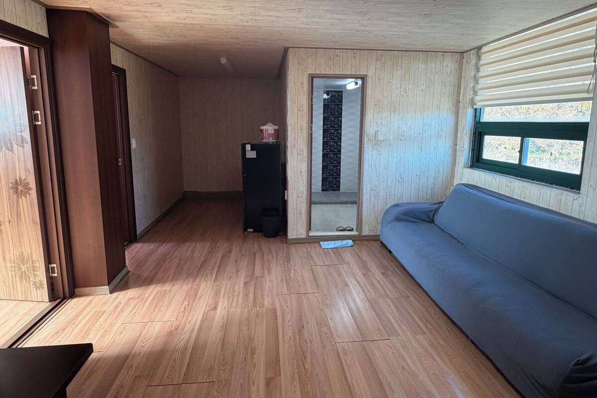 A room with wooden flooring, a sofa, a refrigerator, and a sliding door leading to a balcony.