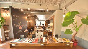A cafe interior with a wooden table displaying magazines, a potted plant, and a chalkboard wall with writing. - Chillulu Coffee & Hostel (Yokohama)
