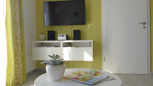 Een kamer met een witte tafel, een kamerplant, een tijdschrift, een televisie aan de muur en een deur.