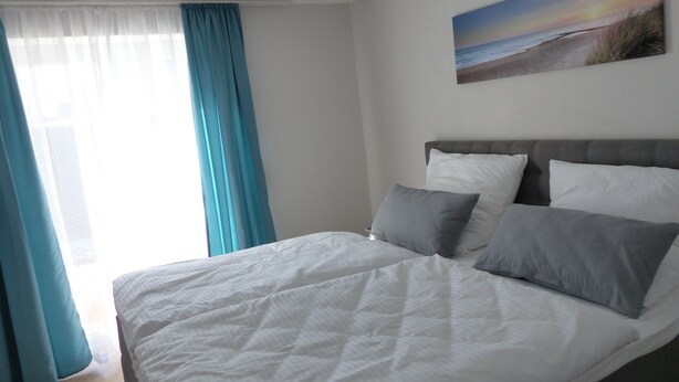Een netjes opgemaakt bed met witte beddengoed en grijze kussens, een grijs bekleed hoofdbord en een schilderij van een strandzicht aan de muur.