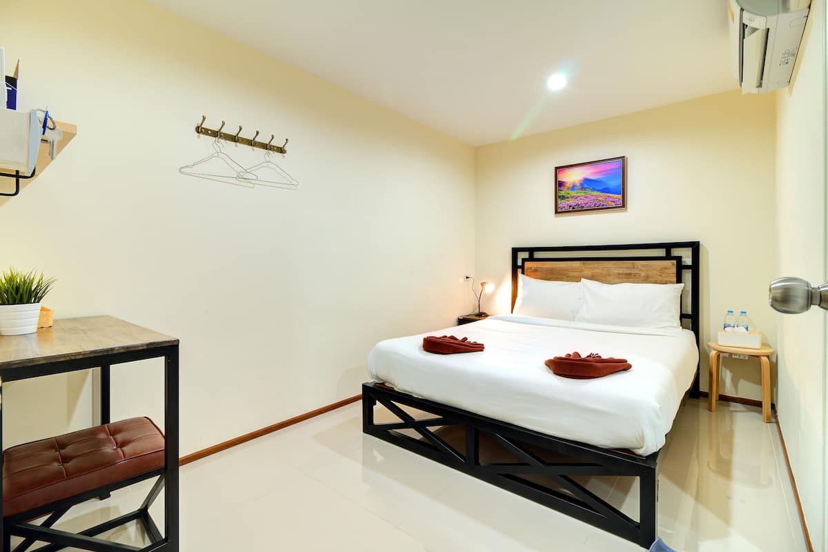 Une chambre d’hôtel comprenant un lit, une petite table avec une chaise, une table de chevet avec une lampe, un porte-manteau fixé au mur et une plante en pot.