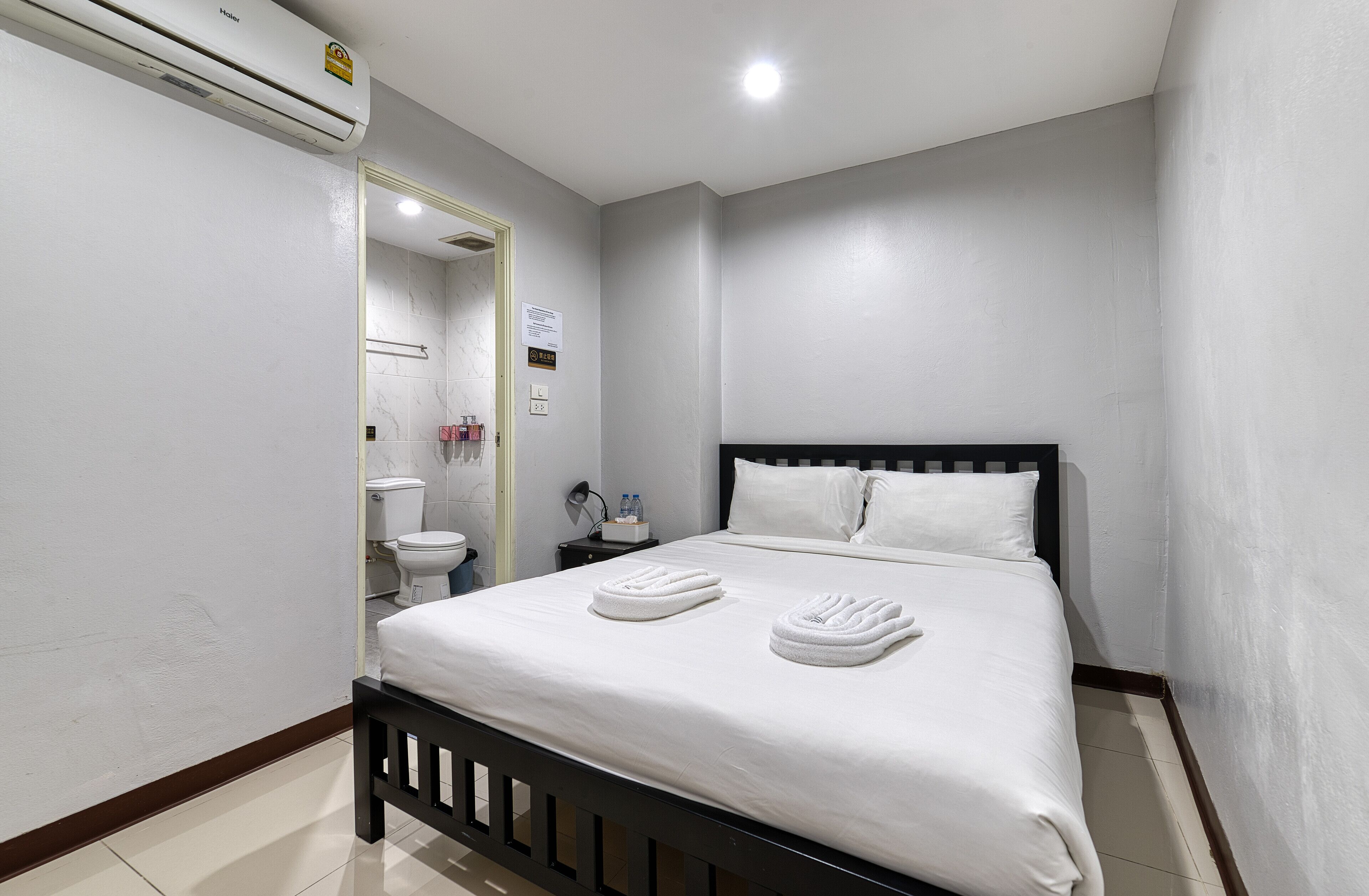 A hotel room with a bed, a bedside table, a nightstand, a mirror, a toilet, and an air conditioning unit.