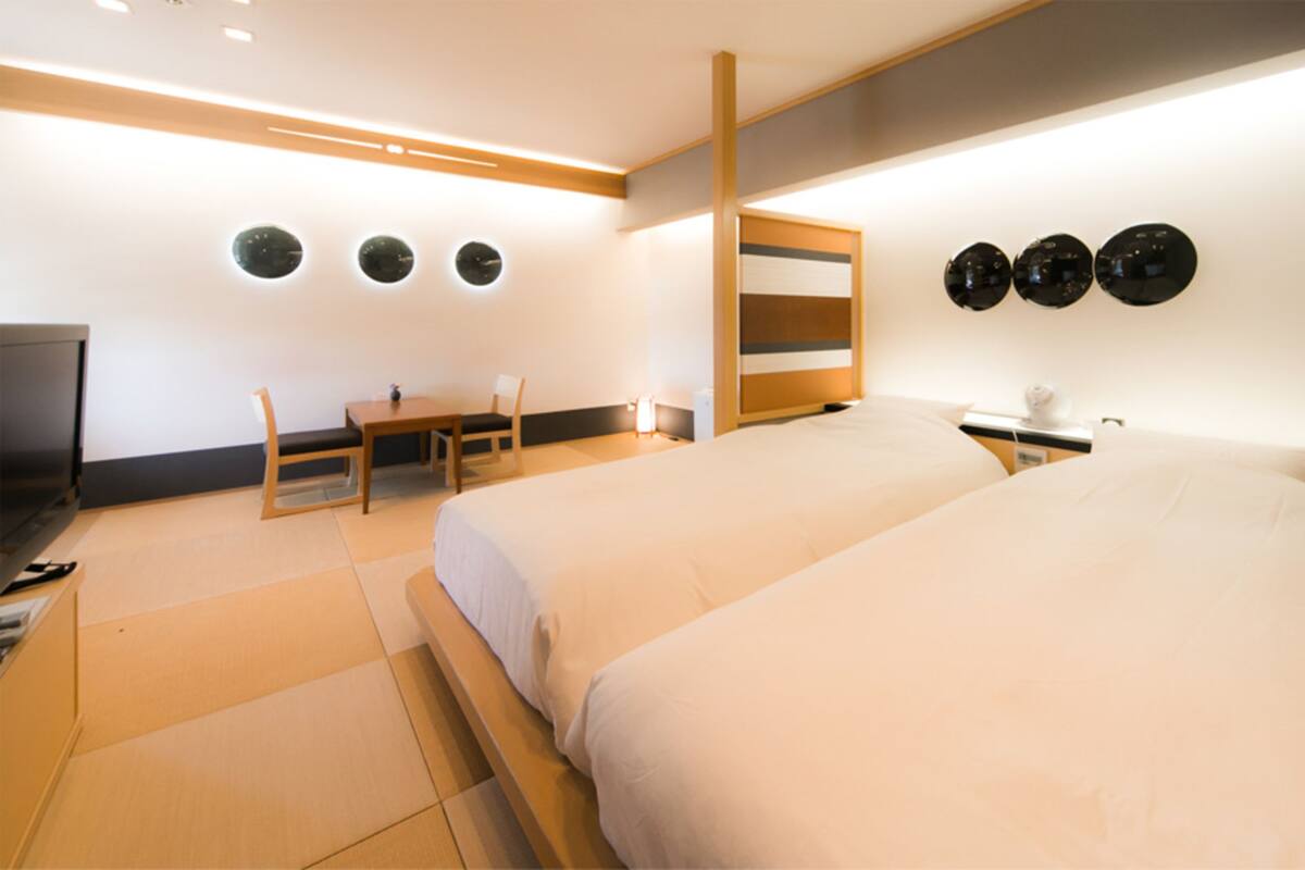 A modern hotel room with two beds, a desk, and a chair. There are three wall-mounted spherical lights and a television on the left.