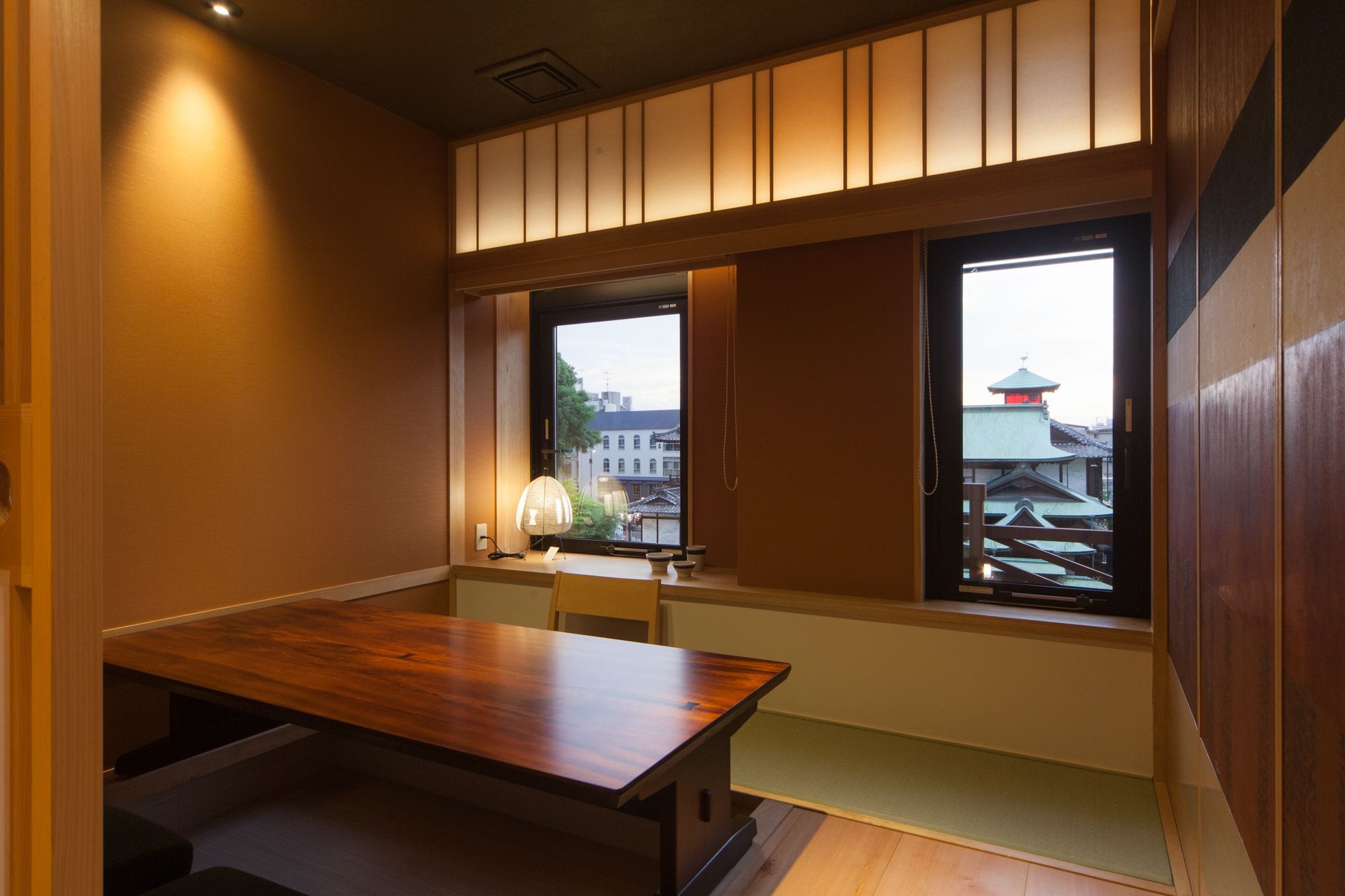 A room with a wooden table and benches, large windows with a view of buildings and a pagoda.