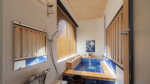 Une petite salle de bain avec une baignoire en bois, une fenêtre avec des stores, un miroir et un espace de douche carrelé de carreaux bleus.