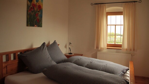 A bedroom with a bed, a nightstand, a window with curtains, a radiator, and a painting on the wall.