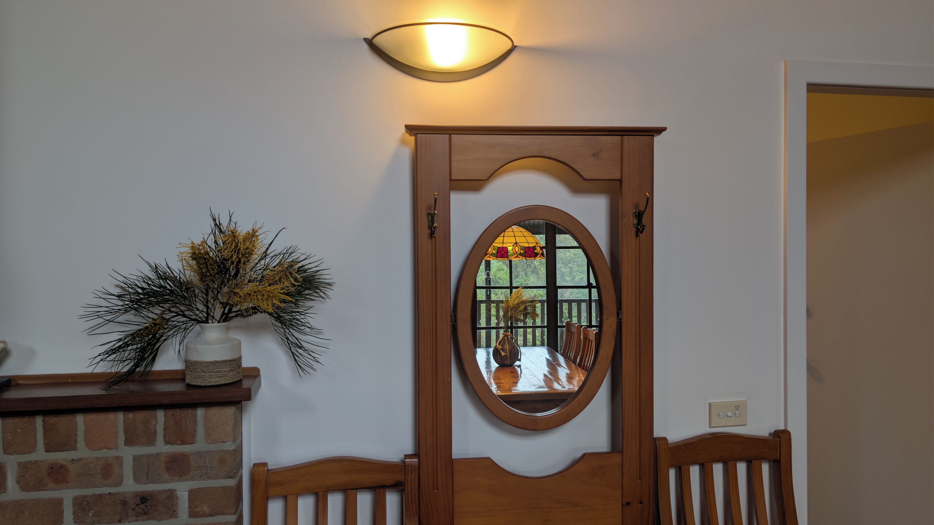 A wooden door with a mirror, a wall-mounted light fixture, and a potted plant with pine branches.