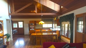 Una sala de estar amplia con techo de madera, un sofá rojo con cojines amarillos, una mesa de comedor de madera con sillas, y una cocina al fondo.