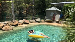 Una persona flota sobre un flotador en una piscina cristalina rodeada de rocas y follaje.