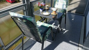 A balcony with a table set for two, chairs, a vase with flowers, and a red car parked outside.