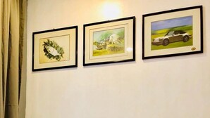 A neatly made bed with white linens in a room with a plain wall. There are three framed pictures above the bed: one of a green wreath, one of a landscape with a house, and one of a car. - Umali Kayo at Megatower Residences 1 (Baguio)