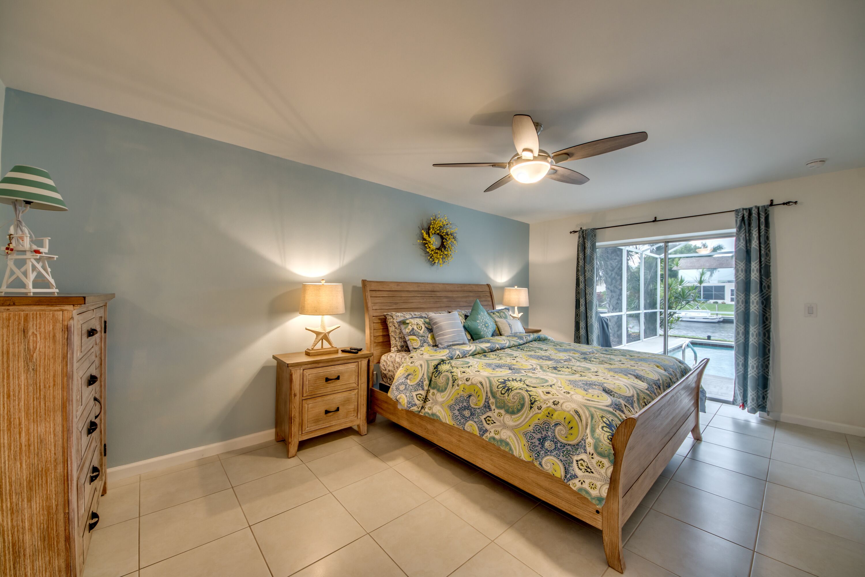 Un dormitorio con cama, mesitas de noche, ventilador de techo, ventana con cortinas y vistas al exterior.
