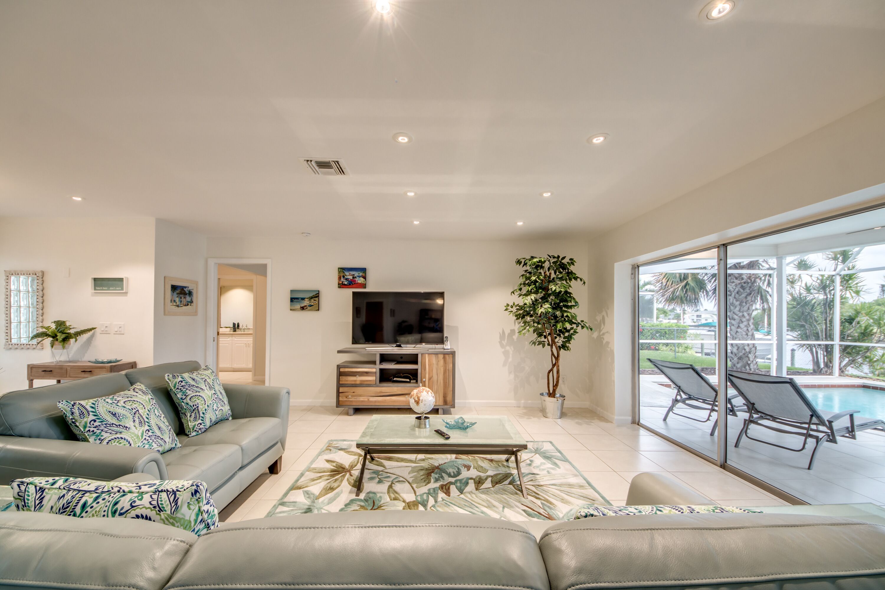 Una sala de estar moderna con un sofá, una mesa de centro y un televisor. Hay un ventanal con vista a una piscina y palmeras.