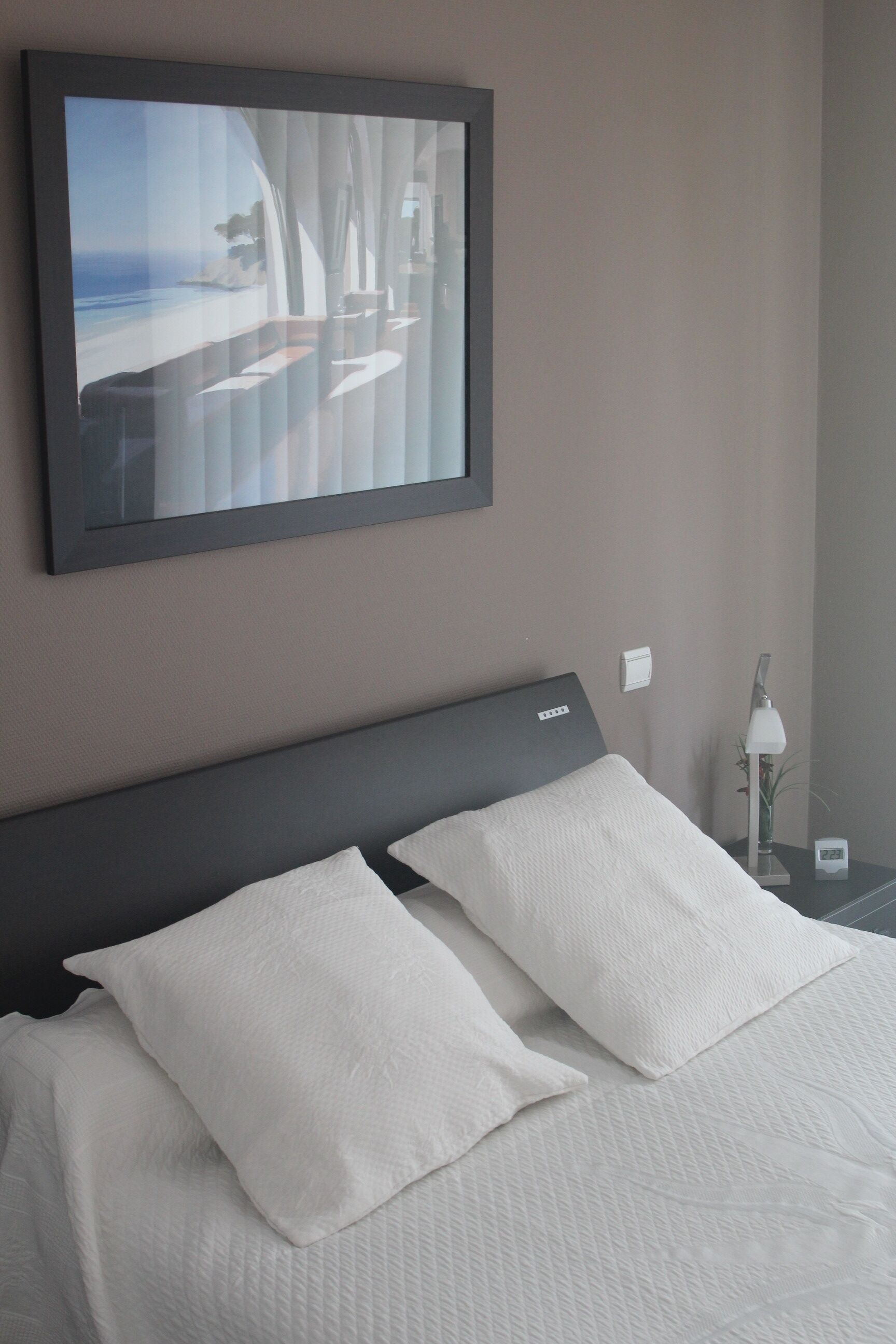 Ein Hotelzimmer mit Bett, weißen Kissen, einem Nachttisch mit Lampe und einem gerahmten Bild an der Wand.