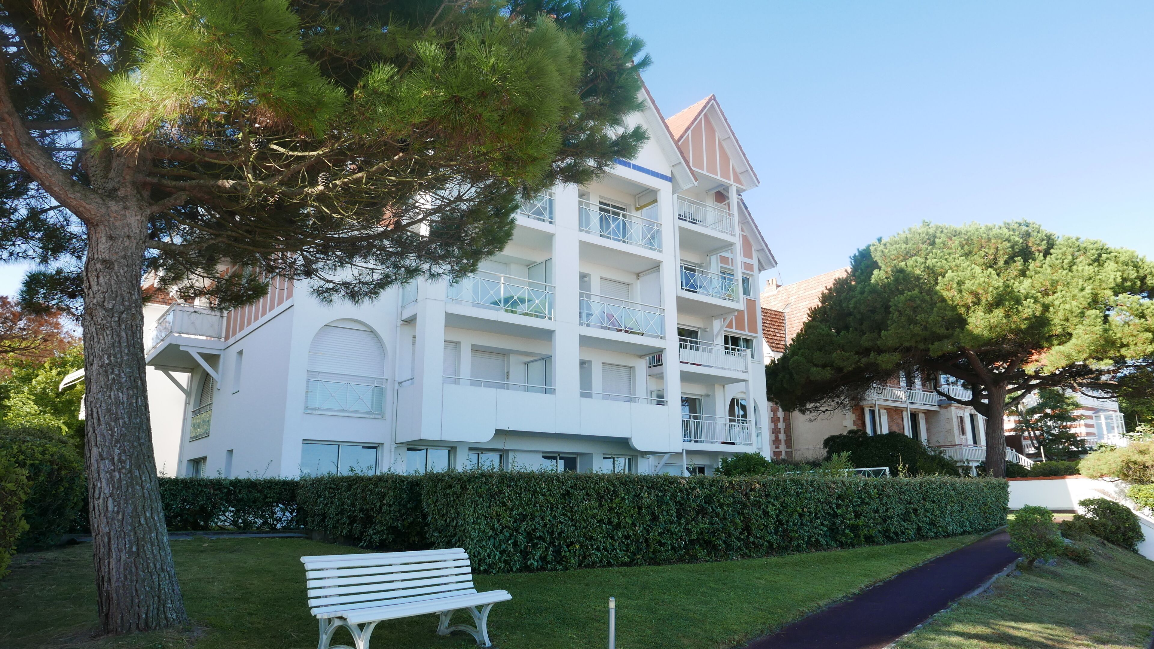 Un immeuble à plusieurs étages doté de balcons, d’un banc et d’une haie au premier plan.