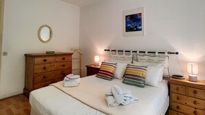 A neatly made bed with pillows, a bedside table with a lamp, a wooden dresser, and a framed picture on the wall. - N&J - MARIN DEBUSSY - Hyper center (Niza)