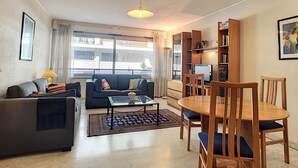 A living room with a sofa, coffee table, and dining table with chairs. There is a bookshelf, a lamp, and a window with curtains. - N&J - MARIN DEBUSSY - Hyper center (Niza)