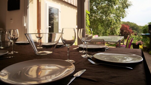 A dining table set with plates, glasses, and cutlery, with a view of a garden and a building through a window.