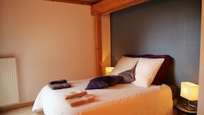 A neatly made bed with a white duvet, several pillows, and folded towels on a wooden floor.