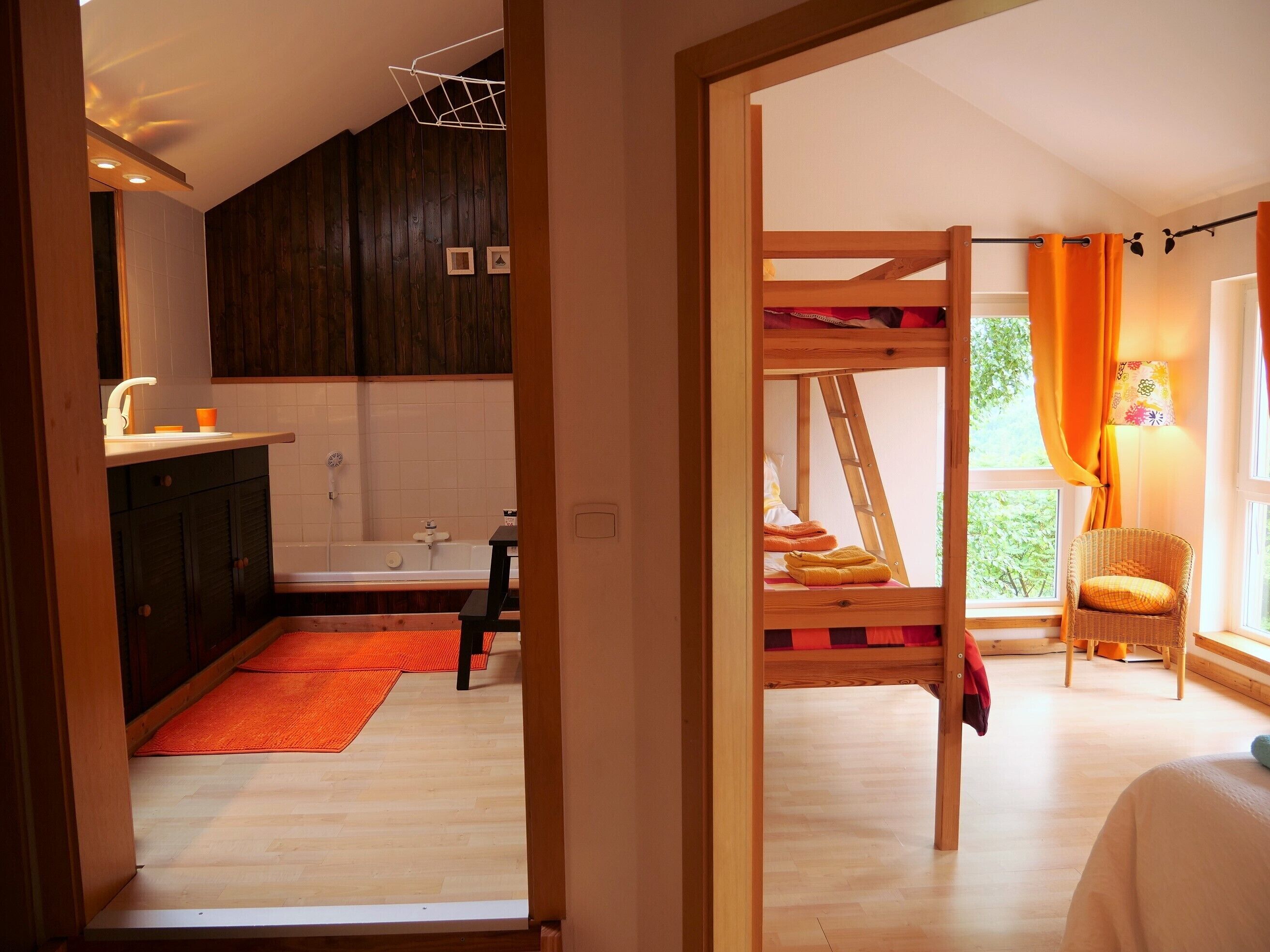 Une pièce avec un lit superposé en bois, une chaise en osier, une fenêtre avec des rideaux orange et une vue sur la verdure à l’extérieur.