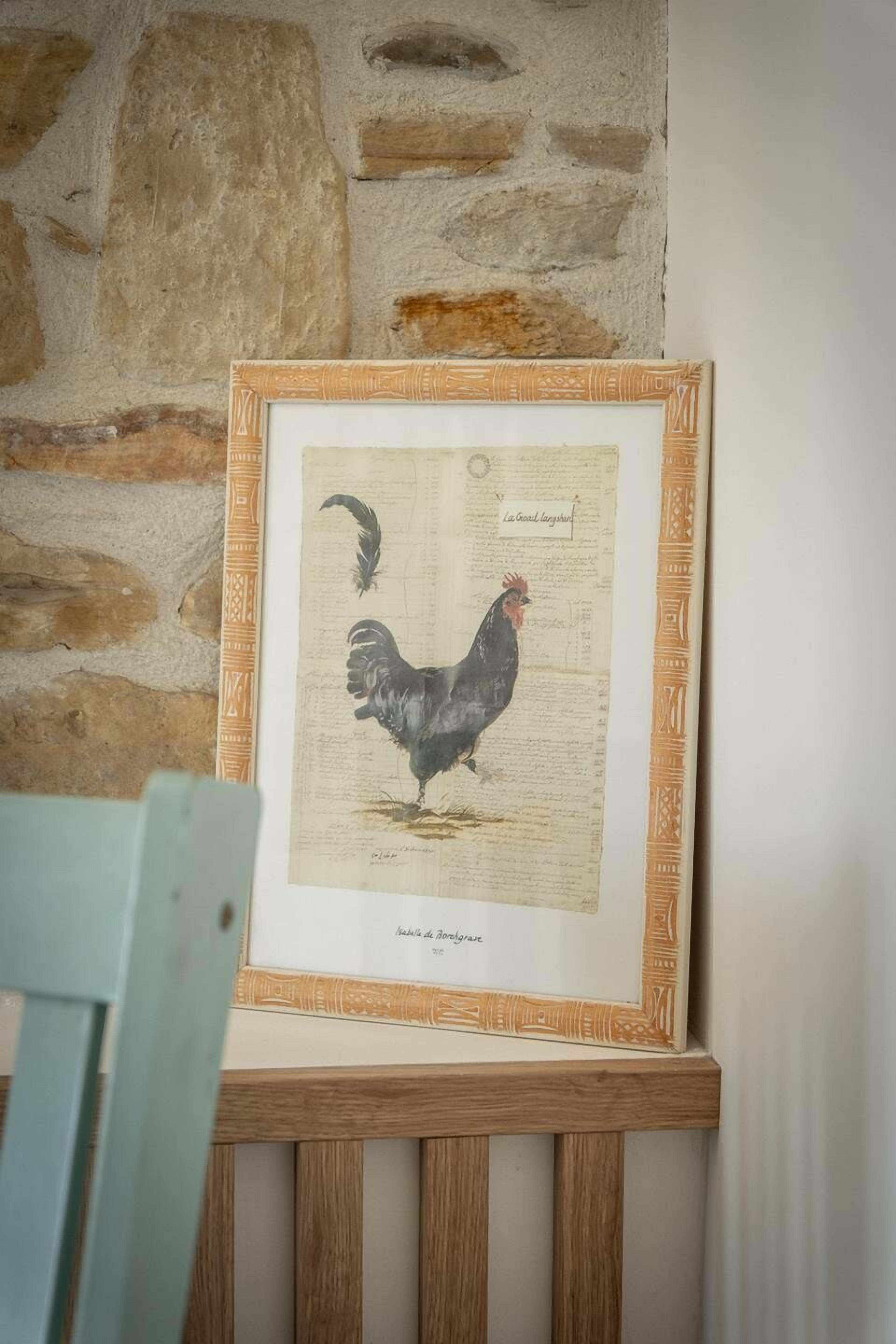 Una obra de arte enmarco con un gallo, sobre un caballete de madera, frente a una pared de piedra.