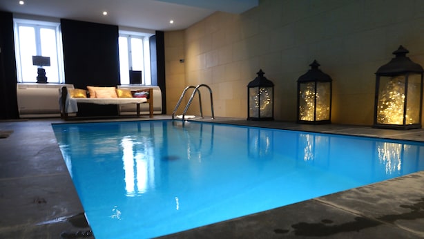 Una piscina interior con agua azul cristalina, un área de descanso con un sofá y una lámpara, y faroles decorativos.