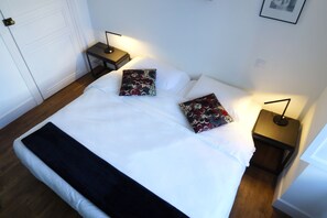 Een netjes opgemaakt bed met witte lakens en twee gestreepte kussens, gelegen tussen twee nachtkastjes met lampen, in een kamer met houten vloer en witte muren.