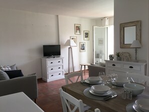 A dining area with a wooden table set for four, white chairs, a white cabinet with a television, and a small round table with a lamp.