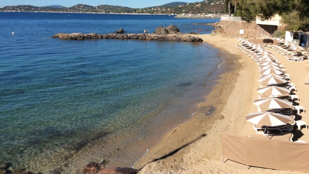 Una spiaggia con acqua cristallina, un molo roccioso e file di ombrelloni.