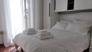 Un letto rifatto con lenzuola e asciugamani bianchi, una finestra con tende trasparenti e un armadio bianco.