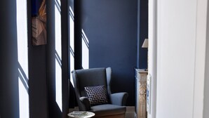 Un pasillo estrecho con pared azul oscuro, suelo de madera y una silla con cojín estampado.