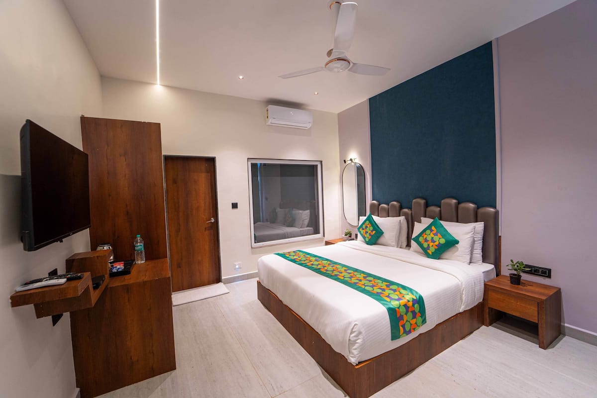 A modern hotel room with a large bed, a TV mounted on a wooden panel, a ceiling fan, and a window with a view of the outside.