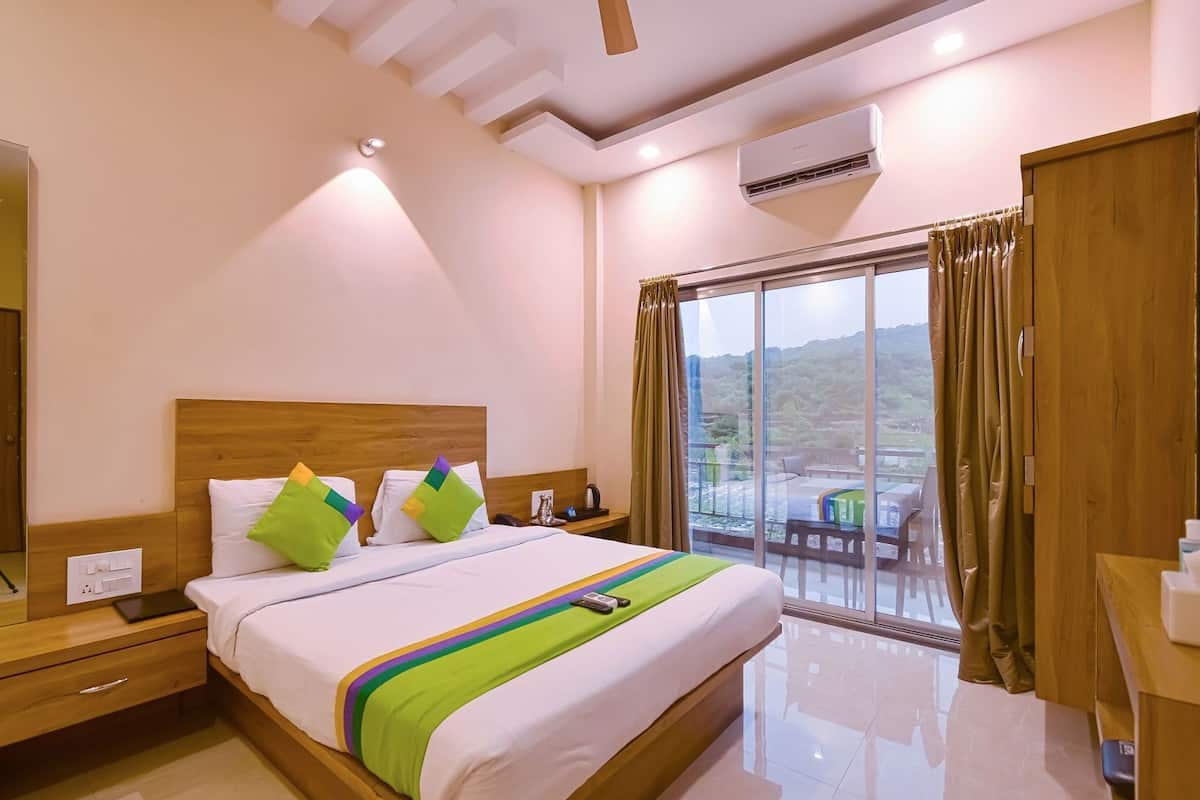A hotel room with a bed, bedside table, nightstand, and a view of the outdoors through a sliding glass door.