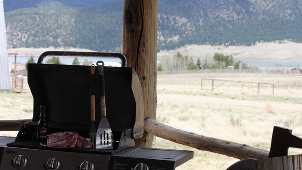 Una parrilla con filetes y una botella de cerveza, frente a un paisaje de montañas y un lago.