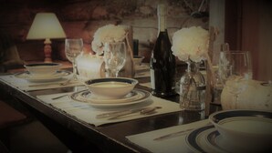 Una mesa de comedor dispuesta con platos, fuentes, cubiertos y una botella de vino, con una vela y flores al fondo.