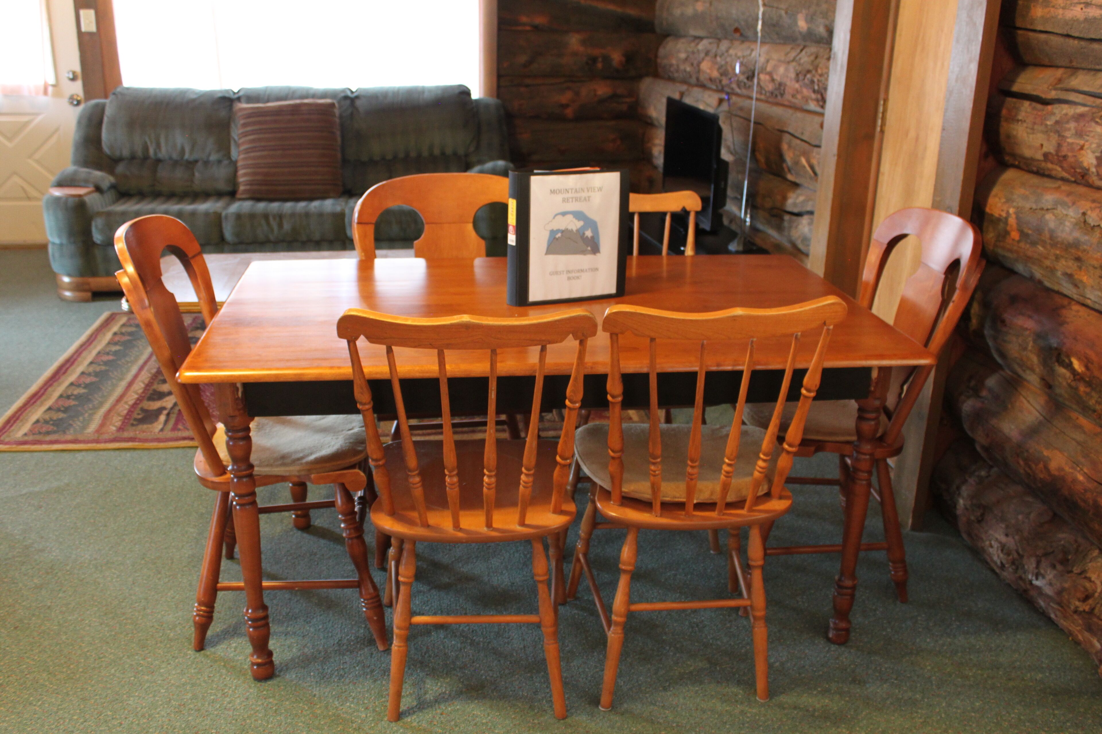 A wooden dining table with four chairs, a book titled "Mountain View Retreat" on the table, and a stone fireplace in the background.