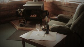 Una habitación acogedora con detalles de madera, una chimenea, una mesa con vasos y una botella, y un sillón cómodo.