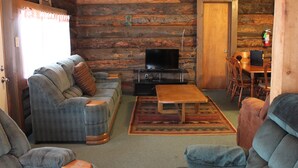 Interior de una cabaña de madera con sofá, mesa de centro, televisor y comedor.