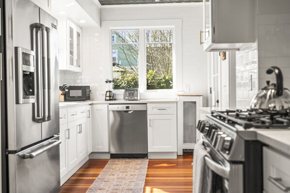 A modern kitchen with stainless steel appliances, white cabinetry, a dishwasher, and a window with a view of greenery.