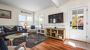 A living room with a sofa, coffee table, and a TV. There are windows, a door, and a bookshelf.