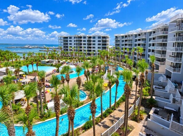 A resort with multiple swimming pools surrounded by palm trees, a marina in the background, and high-rise buildings.