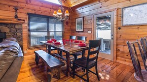 Un interior de madera con una mesa de comedor preparada para cuatro personas, una chimenea de piedra y un ventanal con persianas.
