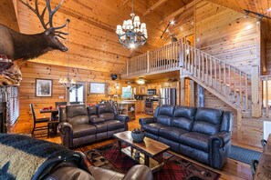 A log cabin interior with a dining area, two leather sofas, a chandelier, and a staircase. - EXPANDED MTN/VALLEY VIEW-500FT NOISY TROUT STOCKED HEMPTOWN CREEK-ARCADE-FIREPIT (Morganton)