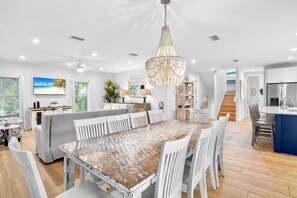 A spacious dining area with a large wooden table surrounded by white chairs, a chandelier, and a view of the beach through the windows.