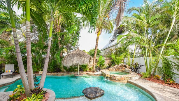 Uma área de piscina tropical com palmeiras, uma estrutura com telhado de palha e uma pequena cascata.