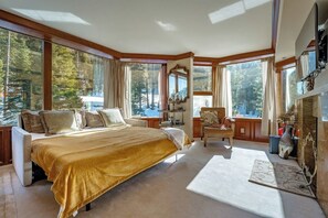 A large bedroom with a king-sized bed, a fireplace, and large windows offering a view of trees.