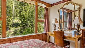 Une pièce dotée d’une grande fenêtre donnant sur la forêt, d’un bureau en bois avec deux lampes et d’un lit recouvert d’une couvre-lit à motifs.