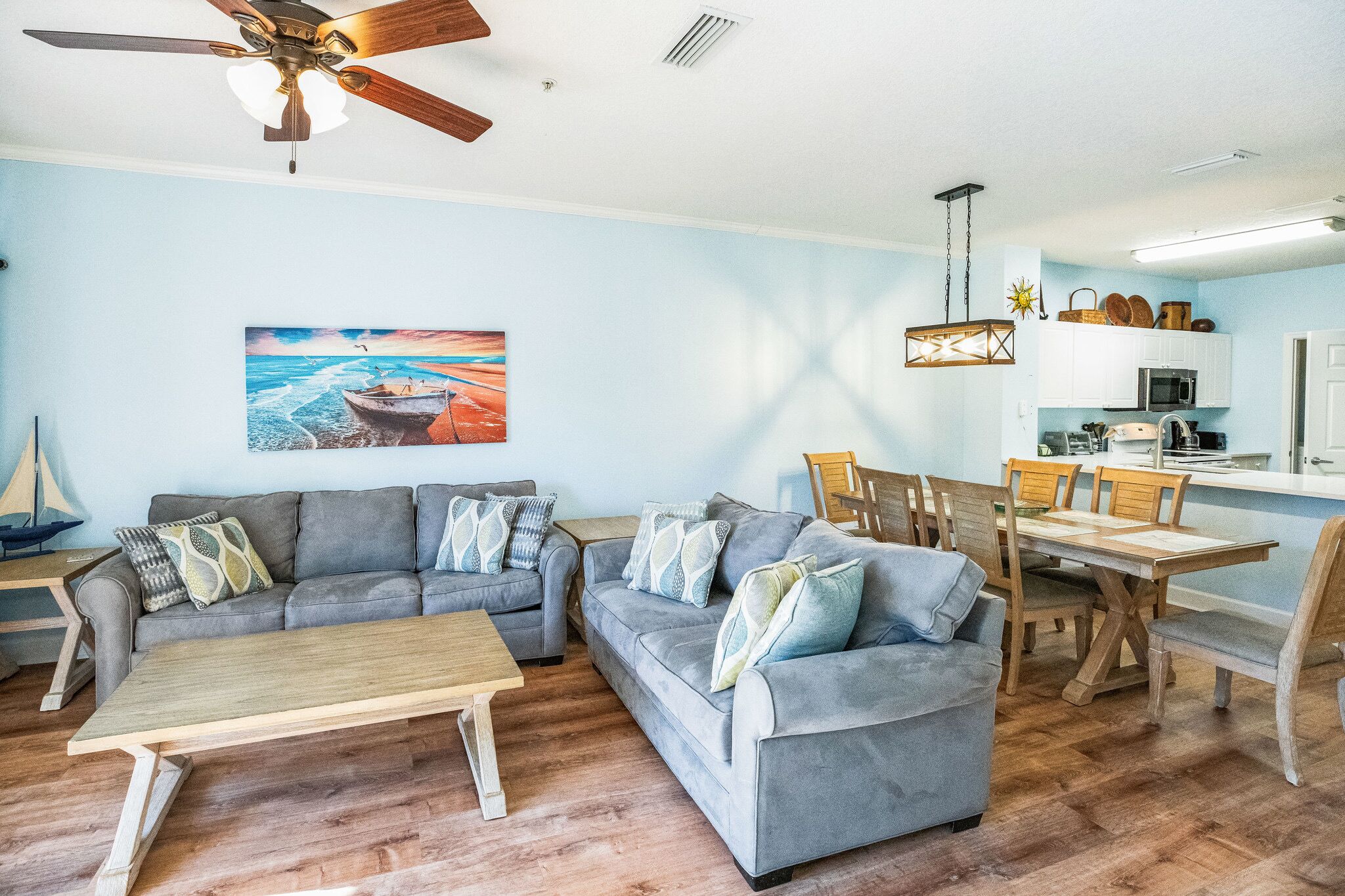 A living room with a gray sofa set, wooden coffee table, and a dining area with a wooden table and chairs. A ceiling fan and a painting of a boat are visible.