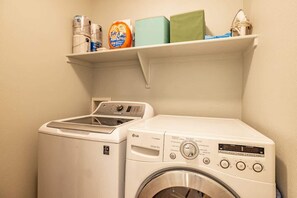 Un espace buanderie comprenant un lave-linge et un sèche-linge, une étagère avec des produits de nettoyage et un robinet fixé au mur.